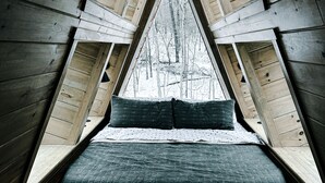 Habitación en ático de forma triangular con vigas de madera, techo inclinado y vista a un bosque nevado a través de una ventana.