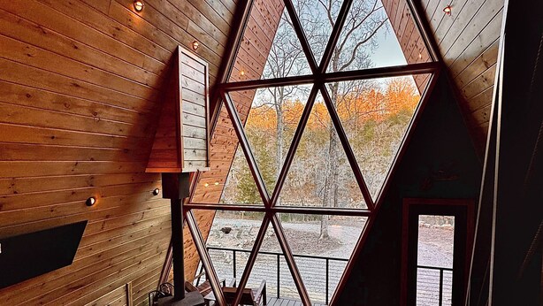 Interior acogedor de una cabaña con un amplio ventanal triangular, paredes de madera, una mesa de madera y un sofá de cuero.