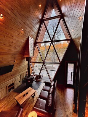 L’intérieur chaleureux d’un chalet avec une grande fenêtre triangulaire, des murs en bois, une table en bois et un canapé en cuir.