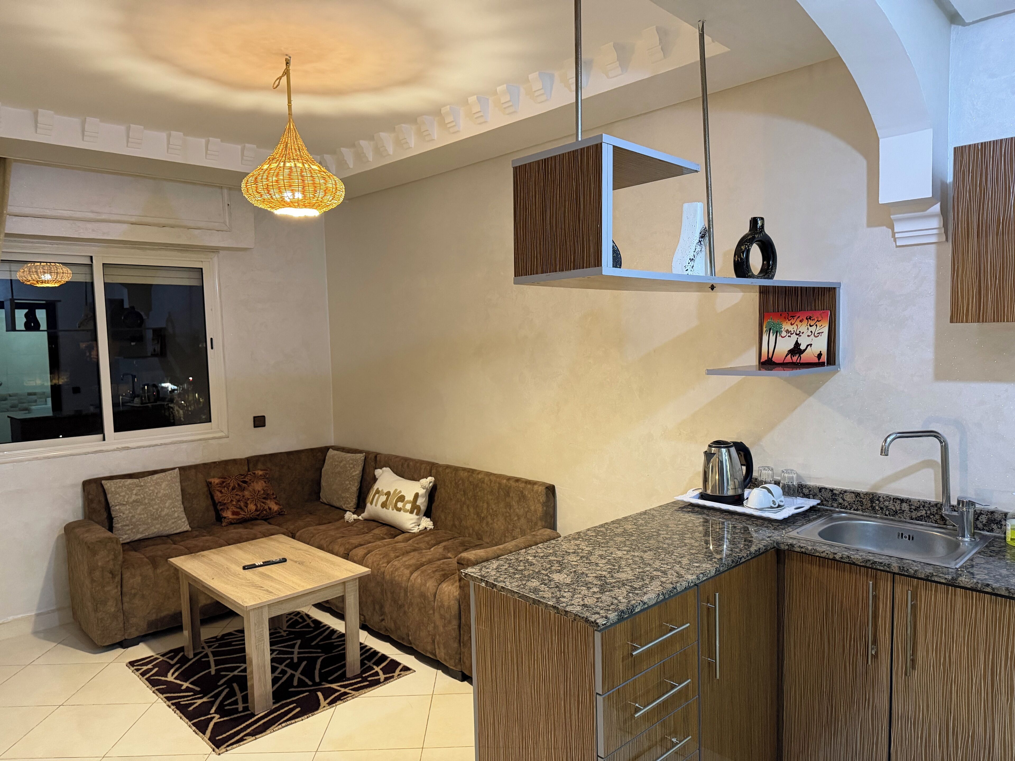 A modern kitchen with a built-in oven, a sink, and a countertop. There is a wooden cabinet with a microwave and a kettle on the counter. A brown sofa with a patterned rug is in the living area.