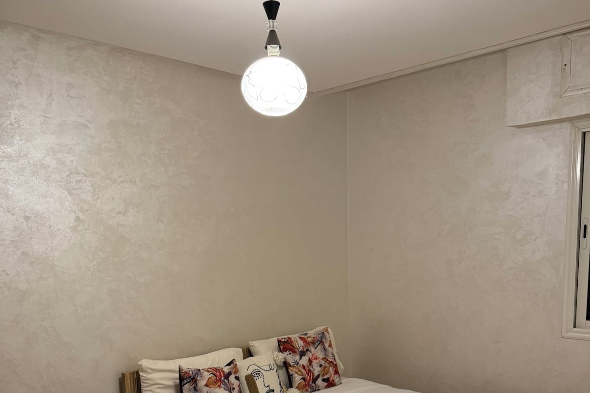 A bedroom with a bed, pillows, a hanging light fixture, a window, and a patterned rug.