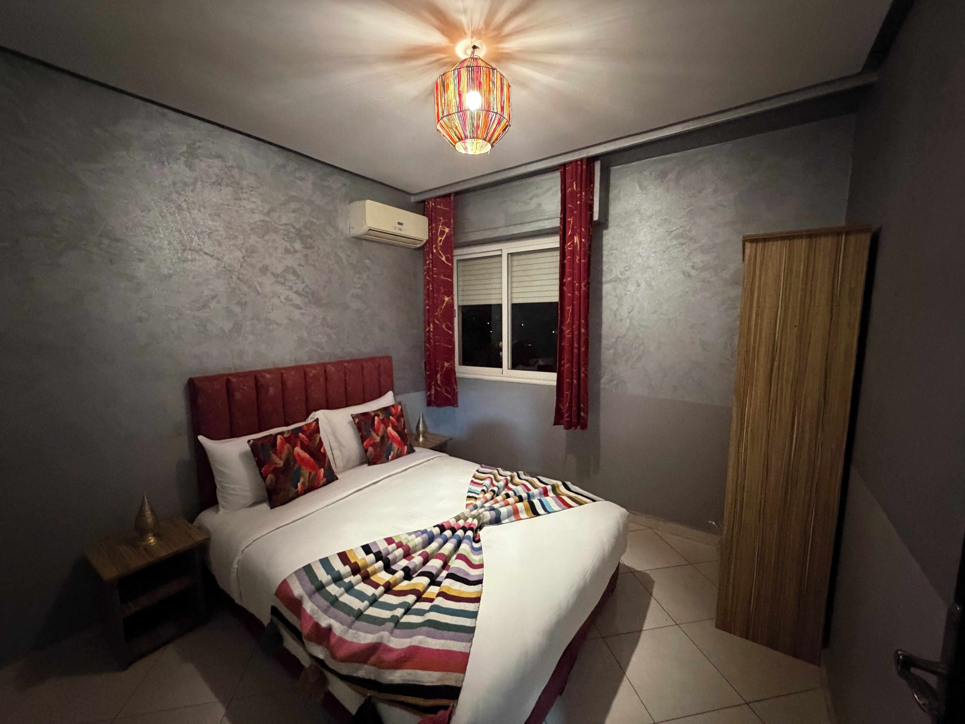 A bedroom with a bed, a nightstand, a window with curtains, an air conditioning unit, and a wooden wardrobe.