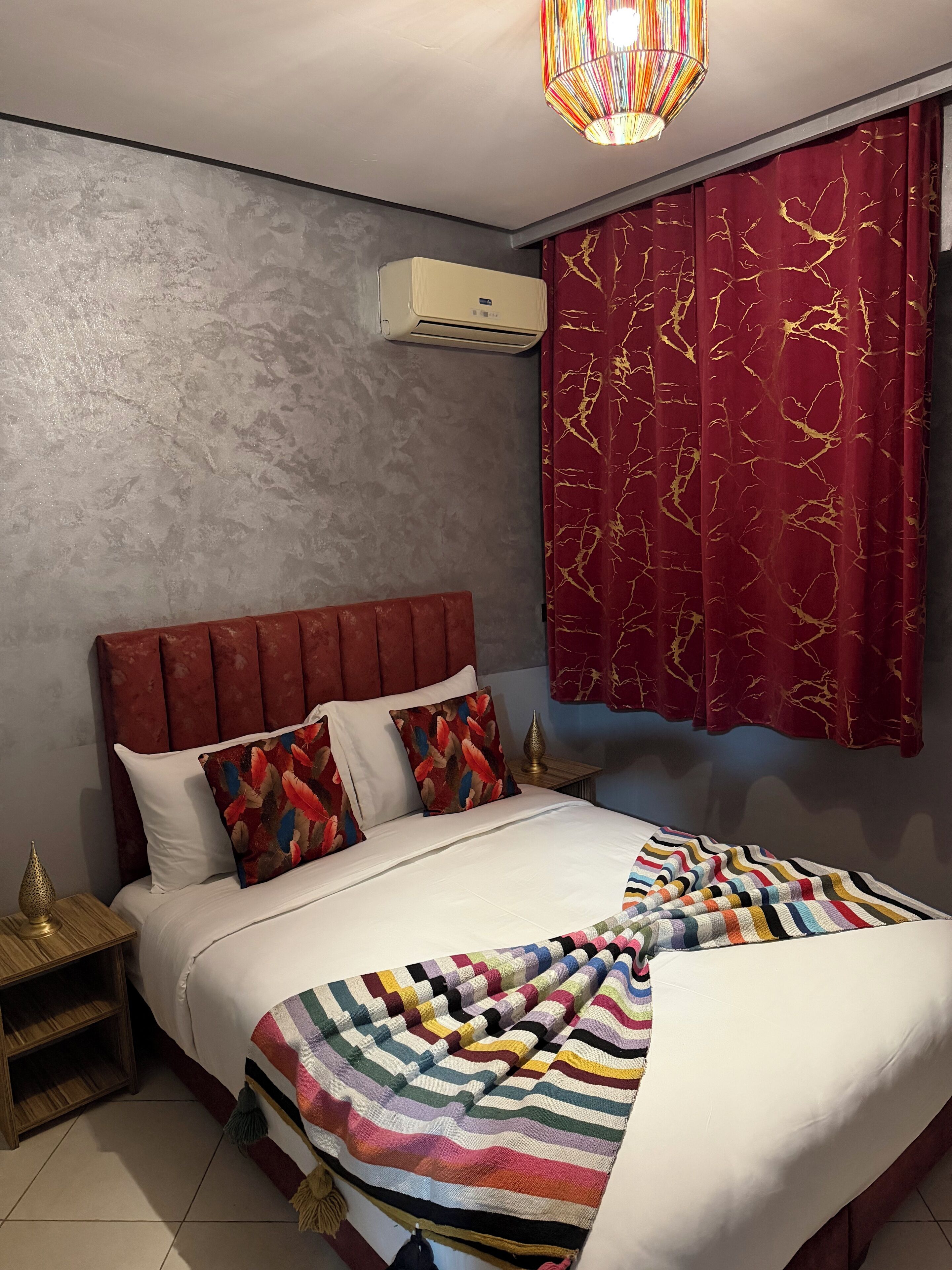 A bedroom with a red headboard, white bedding, a colorful striped blanket, and a red curtain with gold patterns.