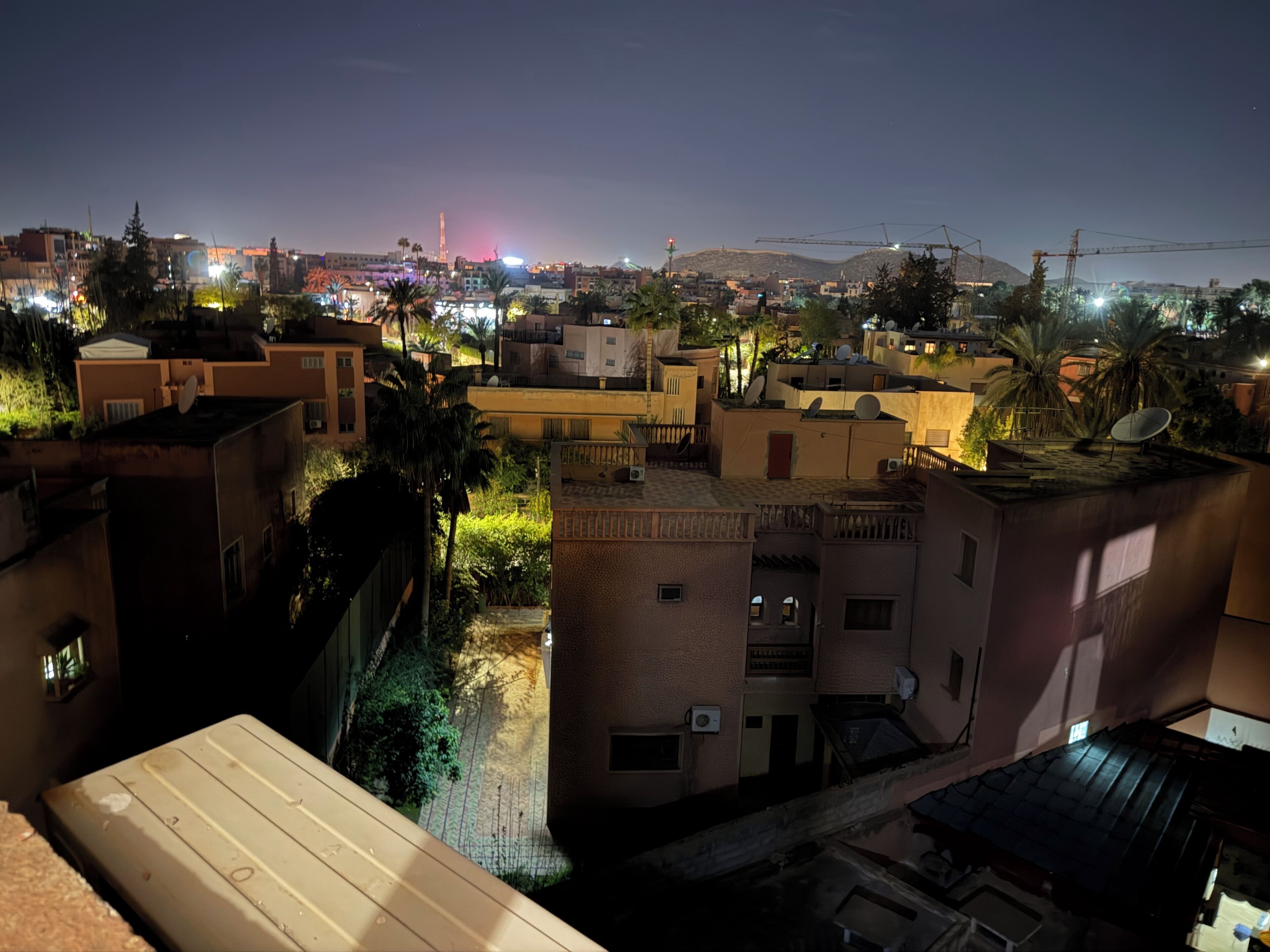 A nighttime view of a residential area with buildings, some with satellite dishes, and a distant cityscape with lights.