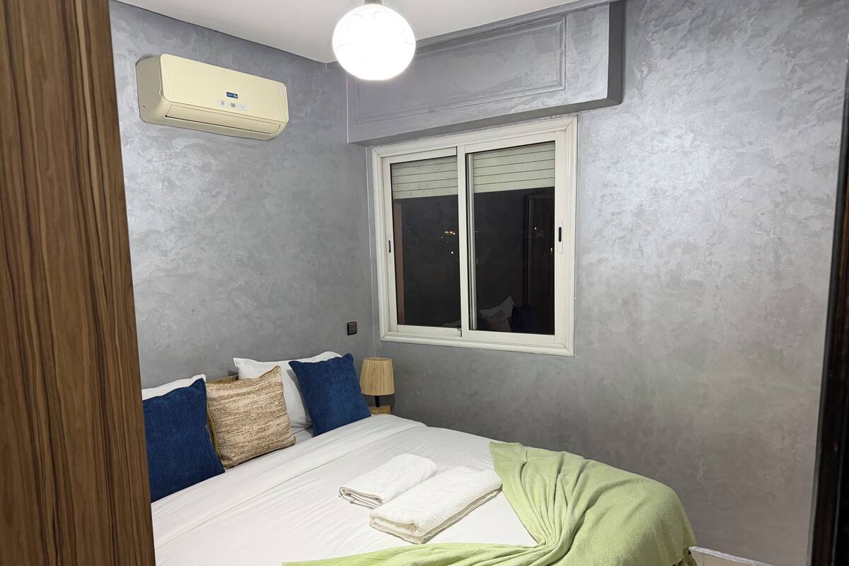 A bedroom with a bed, pillows, a window, and an air conditioning unit.