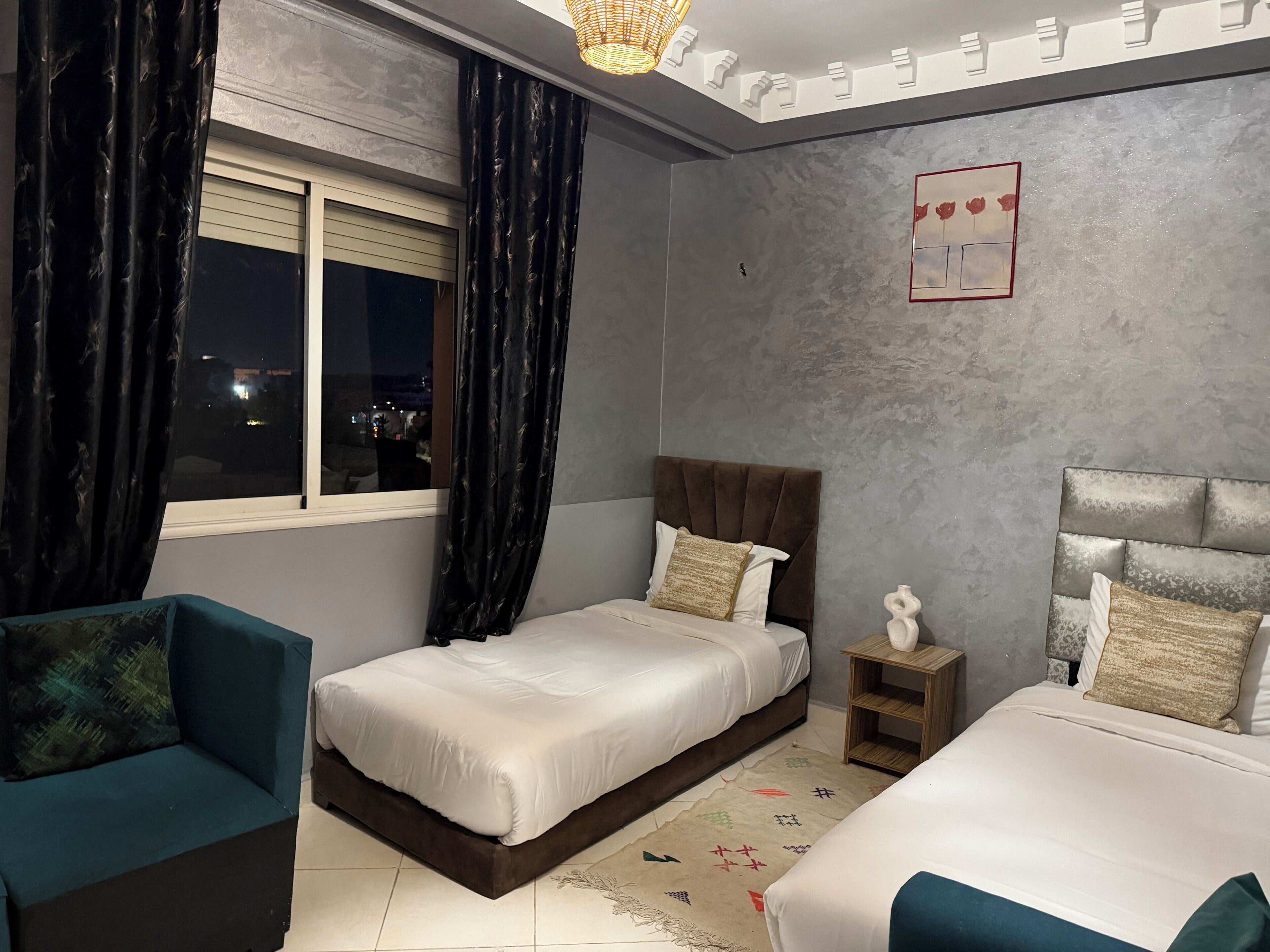 A bedroom with two beds, a window with curtains, a nightstand, and a wall-mounted artwork.
