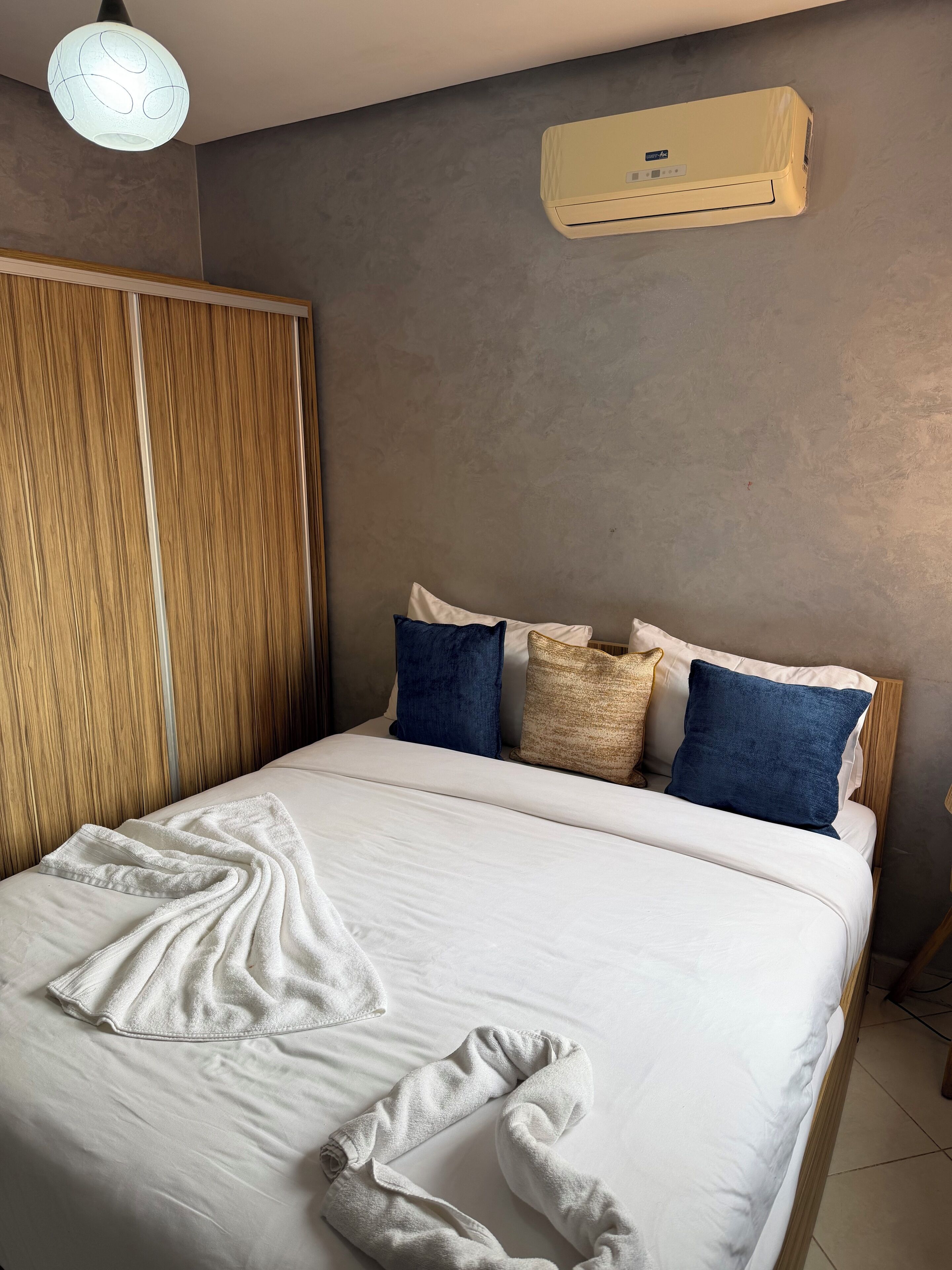 A neatly made bed with white linens and a towel, a wooden wardrobe, and a wall-mounted air conditioner.