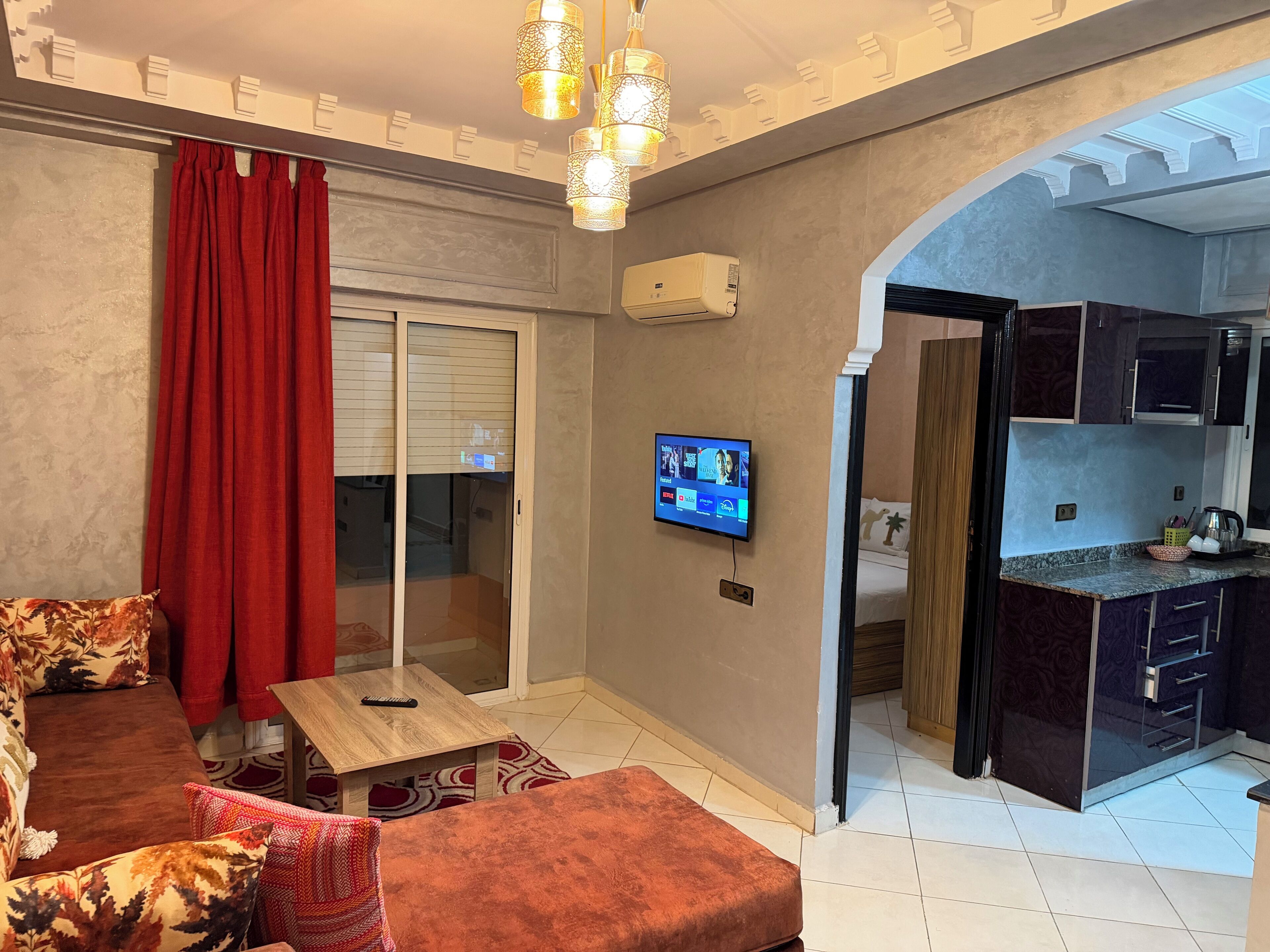 Sala de estar com sofá, mesa de centro e TV fixada na parede. Porta deslizante dá acesso a uma varanda, e cozinha visível através de um arco.