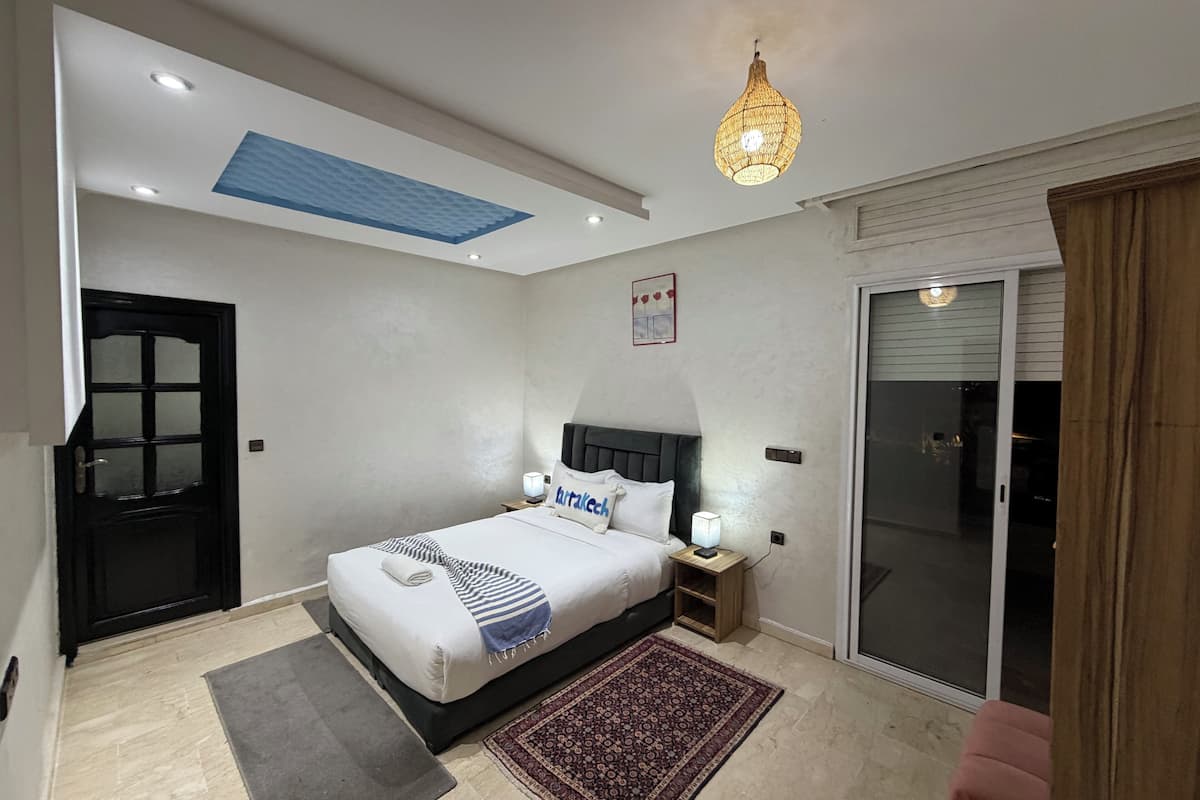 A modern bedroom with a large bed, a ceiling light fixture, a small bedside table, and a sliding glass door leading to a balcony.
