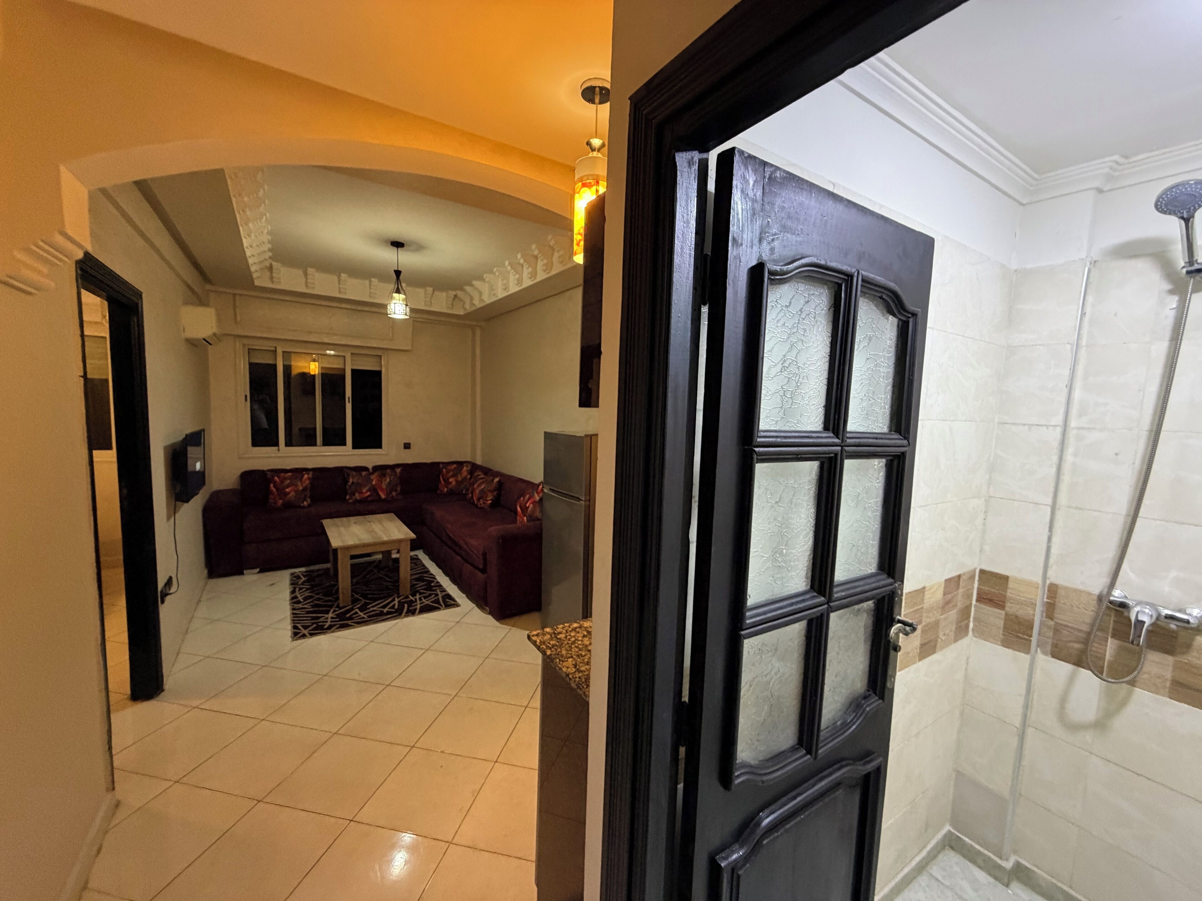 A hotel room with a living area featuring a sofa, a coffee table, and a TV. The room has a tiled floor and a shower with a glass door.