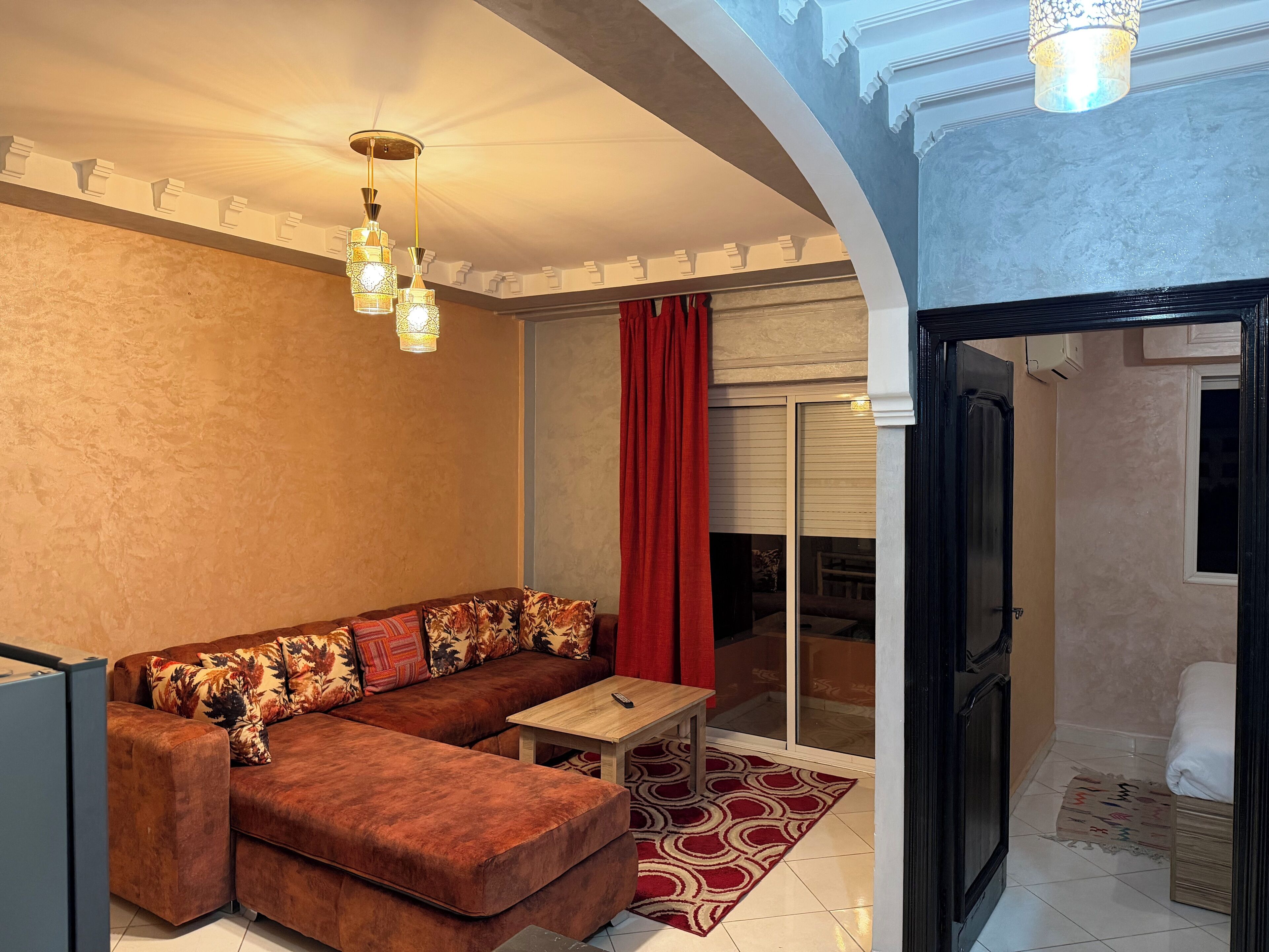 Una sala de estar con un sofá seccional marrón, una mesa de centro de madera y una alfombra roja estampada. Hay tres lámparas colgantes y una cortina roja.