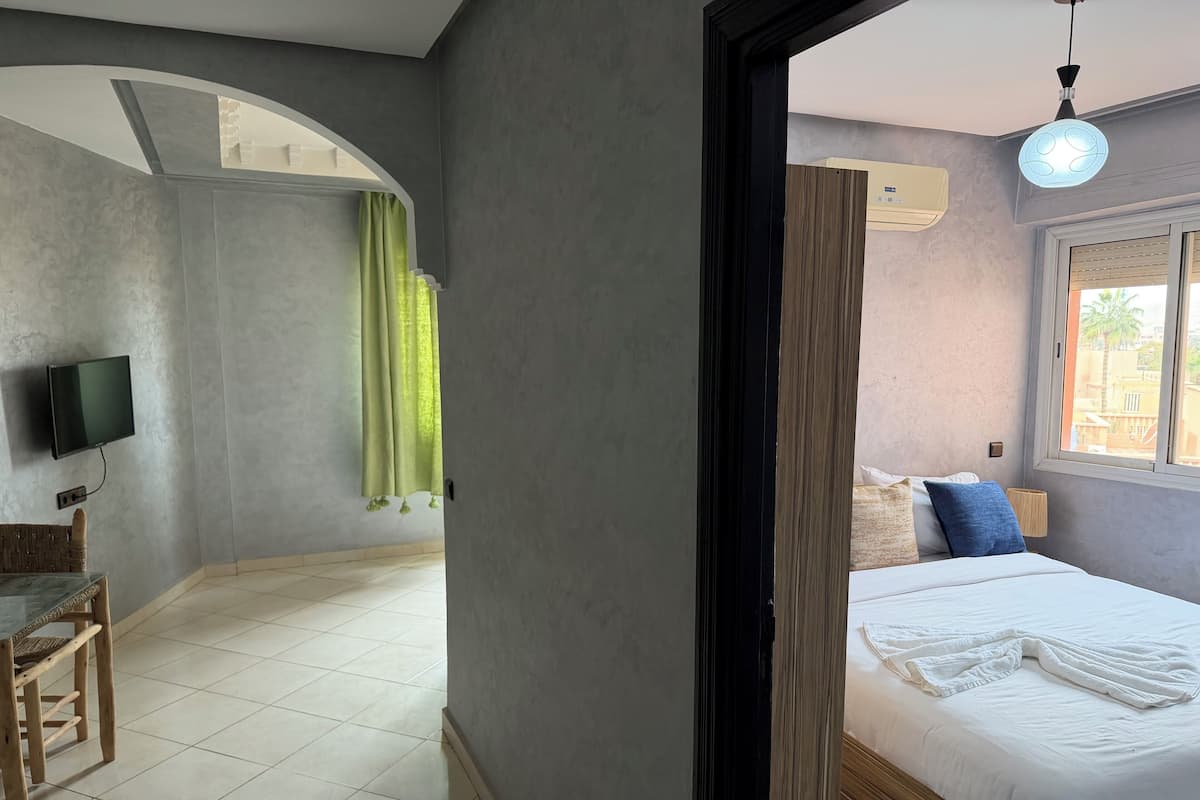 A bedroom with a bed, a TV, a window with blinds, and a view of the outside.
