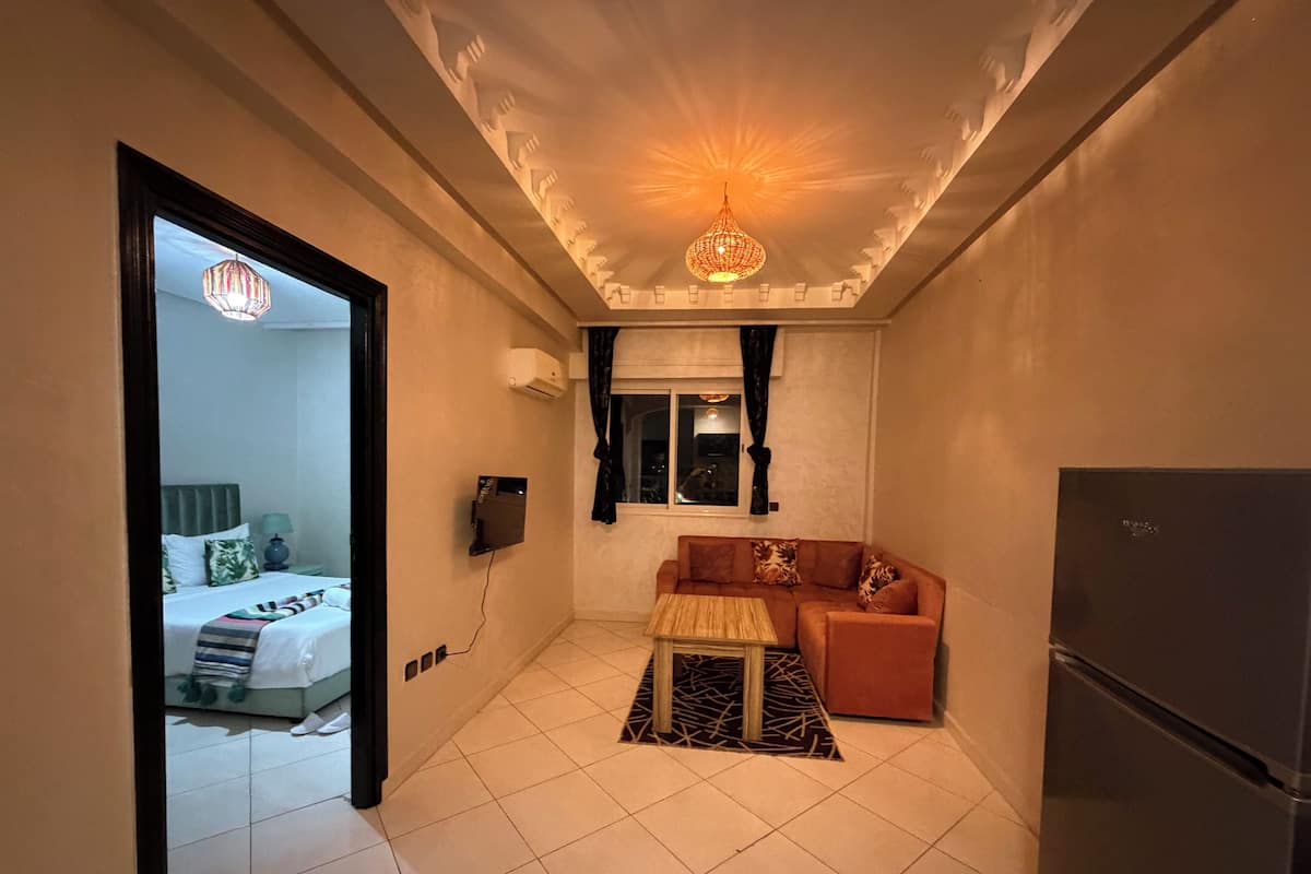 A compact hotel room with a bed, a sofa, a small table, a TV, and a refrigerator.