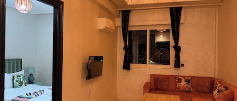 Una habitación con una cama, un sofá, una mesita, un televisor y un ventanal con cortinas.
