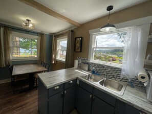 A kitchen with a dining area, two windows with curtains, a sink, and a view of greenery outside. - Scenic Hood Canal View Cottage with AC in charming Union (Union)