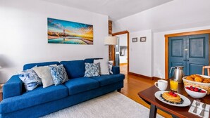 A living room with a blue sofa, a painting of a sunset, a breakfast table with croissants, and a coffee pot. - Bright Upper-Level Retreat - Parking + Deck (Provincetown)