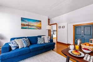 Ein Wohnzimmer mit einem blauen Sofa, einem Gemälde von einem Sonnenuntergang, einem Frühstückstisch mit Croissants und einer Kaffeekanne.
