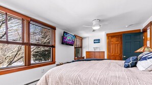 A bedroom with a large bed, a ceiling fan, a TV mounted on the wall, and a view of trees through the windows. - Bright Upper-Level Retreat - Parking + Deck (Provincetown)