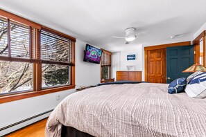 Ein Schlafzimmer mit einem großen Bett, einem Deckenventilator, einem an der Wand befestigten Fernseher und Blick auf Bäume durch die Fenster.