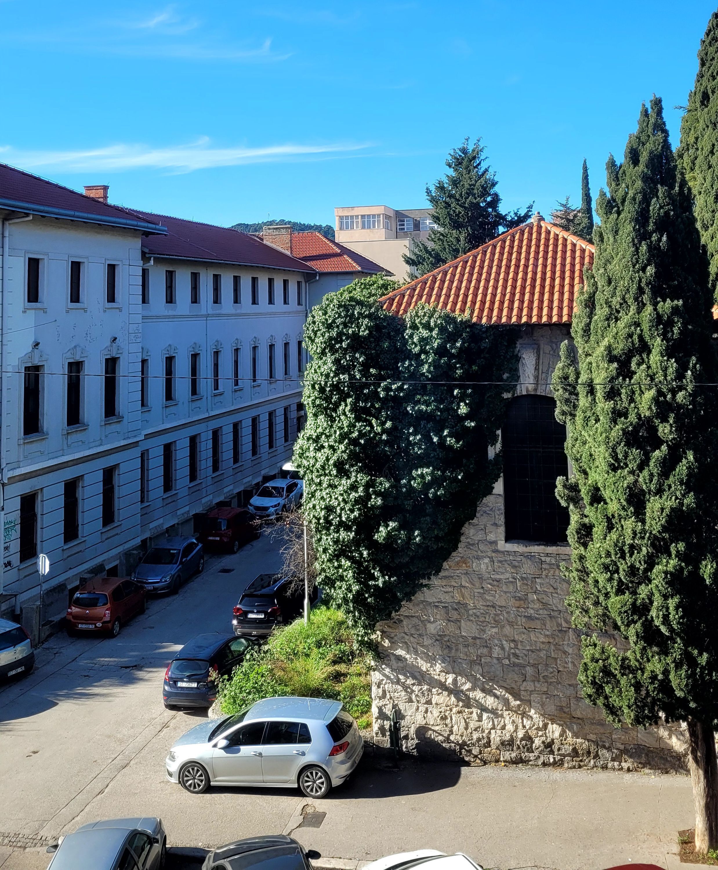 Straßenansicht mit parkenden Autos, einem Steinhaus mit Ziegeldach und einer Reihe weißer Gebäude mit Rundbogenfenstern.