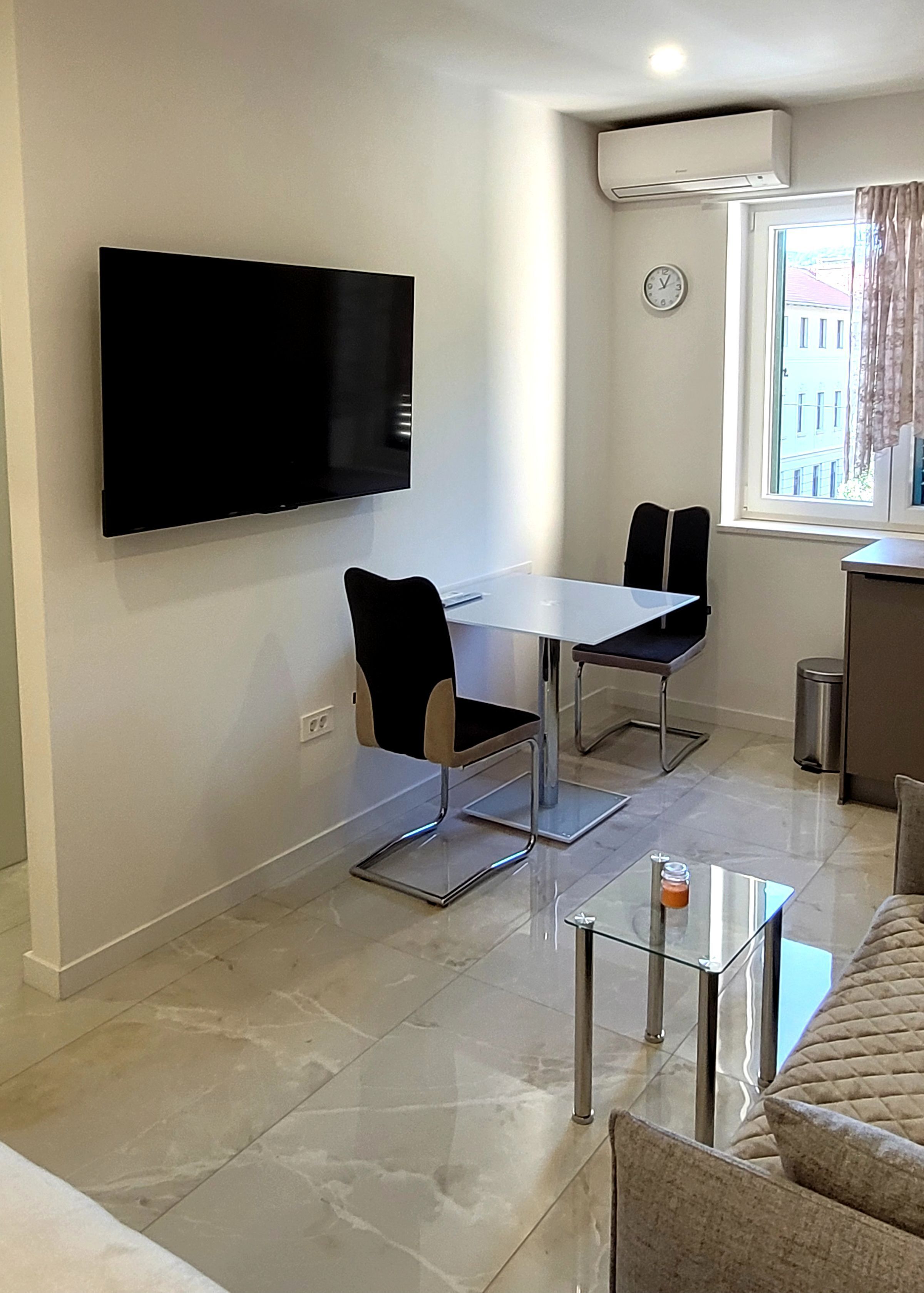 Una sala de estar moderna con una televisión de pantalla plana montada en la pared, una mesa de comedor con dos sillas, una mesa de centro de vidrio y un sofá.