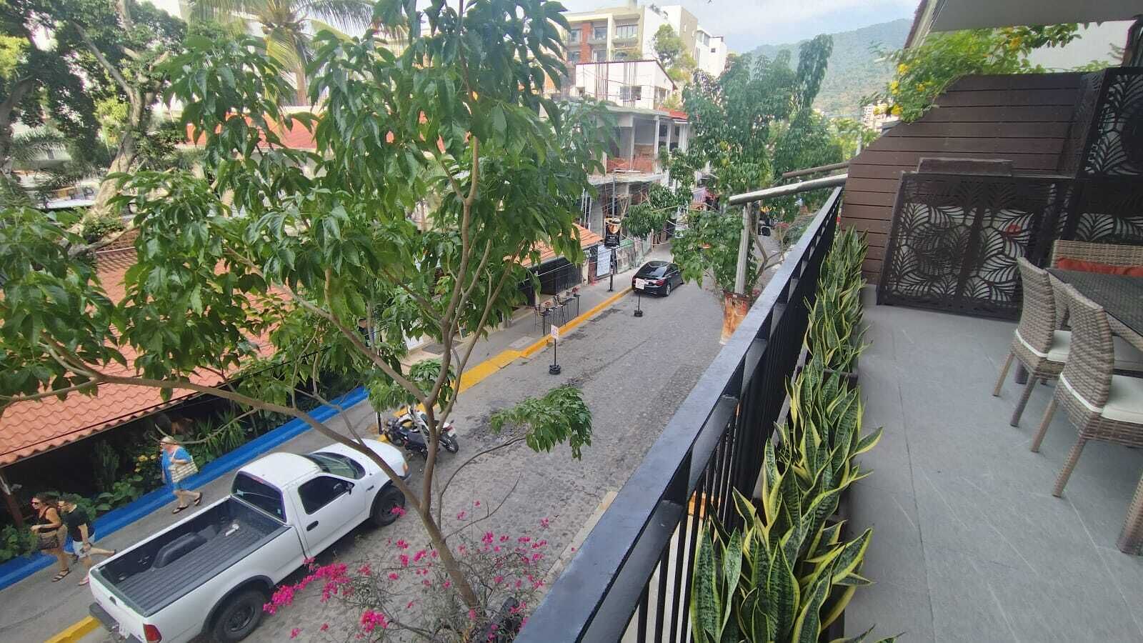A balcony with a view of a street, a white pickup truck parked on the street, and a small garden area with pink flowers.