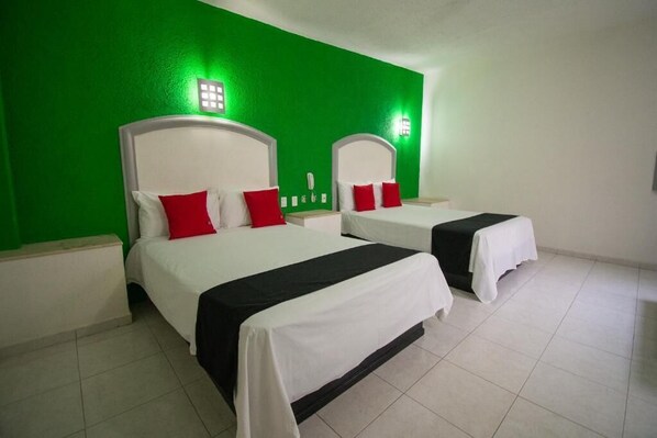 A hotel room with two beds, each with white headboards and red pillows. The walls are green, and there are two wall-mounted light fixtures.