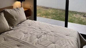 Ein Schlafzimmer mit Bett, Nachttisch, Lampe und Blick auf eine Grünfläche durch große Fenster.