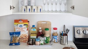 Una cocina con armarios blancos, una estufa y varios utensilios como una sartén, platos, tazas y una tabla de cortar con cubiertos.