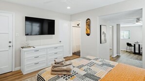 A modern bedroom with a large bed, a TV mounted on the wall, a ceiling fan, and a dining area visible through an open door. - Spacious Suite: Minutes to Everything! (San Diego)