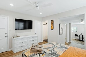 A modern bedroom with a large bed, a TV mounted on the wall, a ceiling fan, and a dining area visible through an open door. - Spacious Suite: Minutes to Everything! (San Diego)