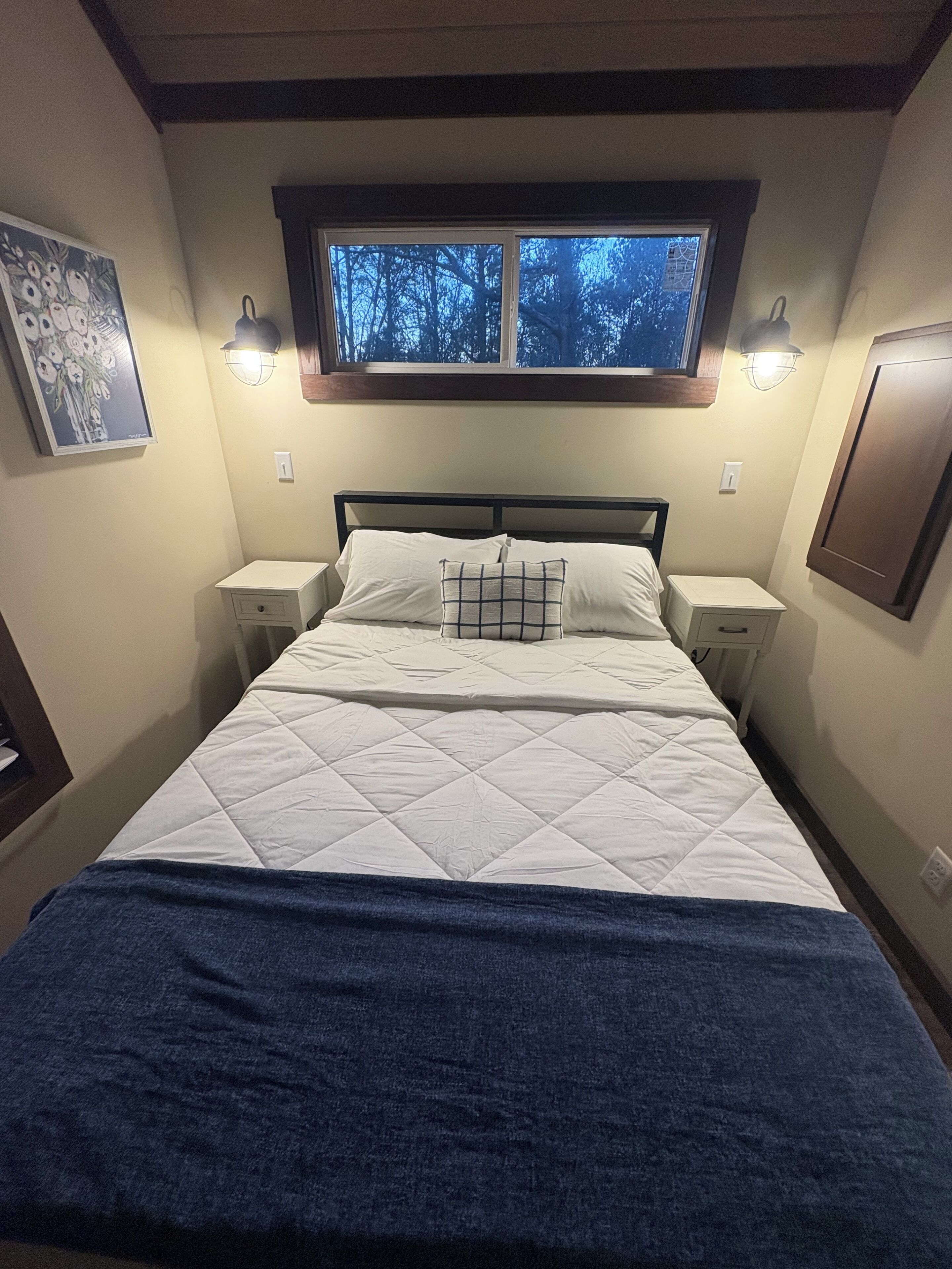 Una cama bien hecha con un edredón blanco acolchado, una manta azul y una almohada de cuadros. Hay dos mesitas de noche con lámparas, una ventana sobre la cama y una lámpara de pared.