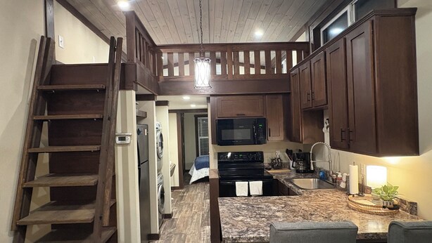 A kitchen with wooden cabinets, a stainless steel refrigerator, a microwave, and a sink. There is a wooden staircase on the left and a dining area with a marble countertop and chairs.