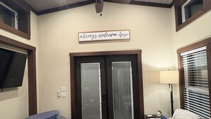 Una sala de estar con un ventilador de techo, dos puertas con persianas, un cartel que dice "siempre bienvenidos", un televisor montado en la pared y una lámpara sobre una mesita auxiliar.