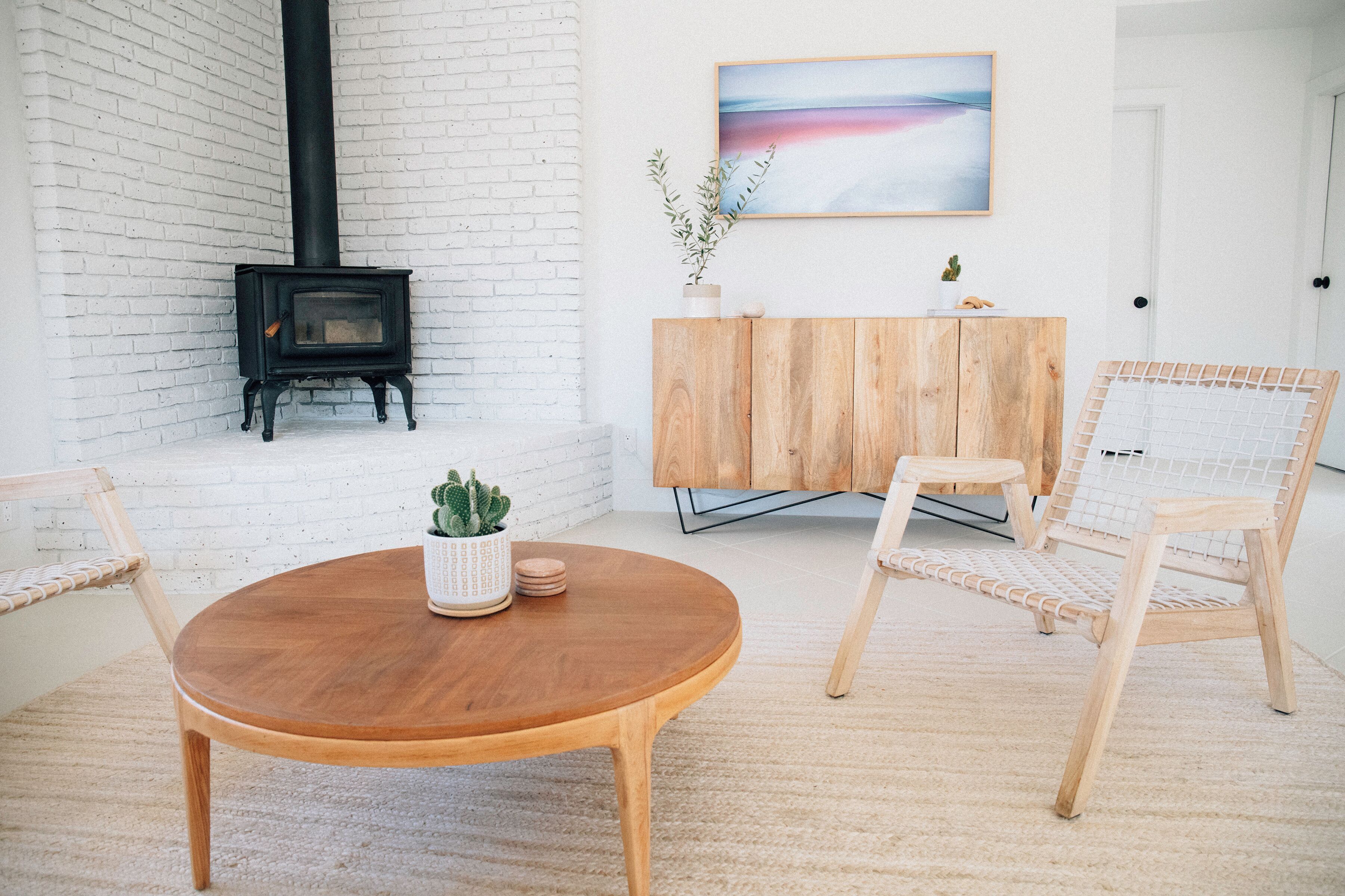Ein Zimmer mit einem runden Holztisch, zwei Korbstühlen, einer weißen Backsteinwand, einem schwarzen Kaminofen, einem Holzschrank und einem Gemälde an der Wand.