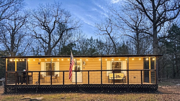 A wooden cabin with a deck, a flag, and a view of trees. - Walk to Lake • 🔥Fire Pit • Pets Welcome 🐾.             + golf ⛳️ (Canadian)