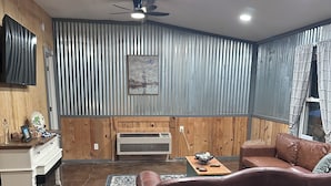 A living room with a brown leather sofa, a wooden fireplace, a ceiling fan, and a large window with curtains. - Walk to Lake • 🔥Fire Pit • Pets Welcome 🐾.             + golf ⛳️ (Canadian)