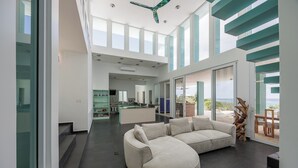 A spacious living area with a large L-shaped sectional sofa, a kitchen in the background, and a view of the outdoors through large windows. - Casa Sea Glass 5 bedroom home waterfront with Pool (Roatan)