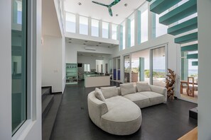 A spacious living area with a large L-shaped sectional sofa, a kitchen in the background, and a view of the outdoors through large windows. - Casa Sea Glass 5 bedroom home waterfront with Pool (Roatan)