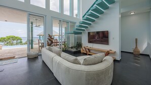 A modern living room with a large window, a staircase, and a flat-screen TV. - Casa Sea Glass 5 bedroom home waterfront with Pool (Roatan)