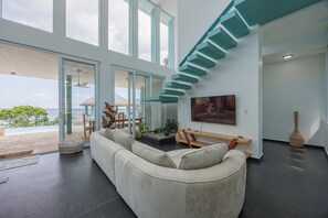 A modern living room with a large window, a staircase, and a flat-screen TV. - Casa Sea Glass 5 bedroom home waterfront with Pool (Roatan)