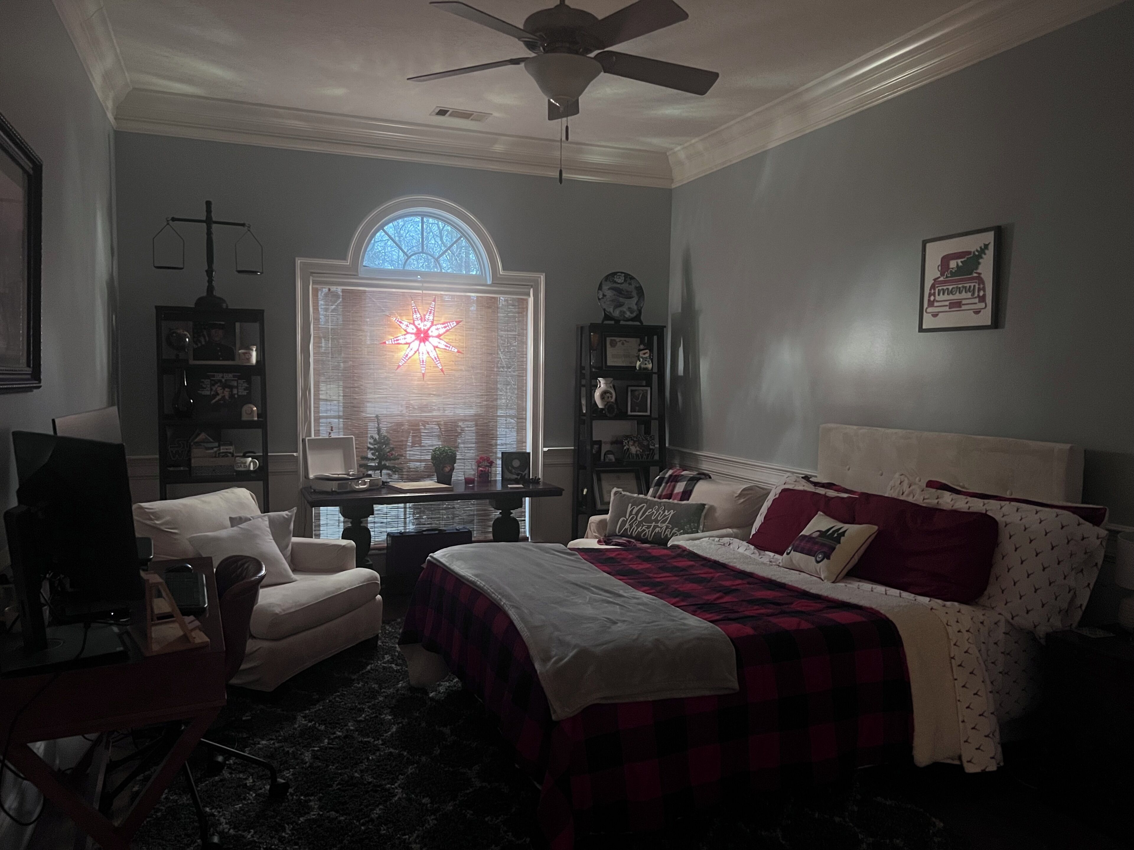 A bedroom with a bed, a desk, a chair, a window with blinds, a ceiling fan, and a wall with a framed picture.