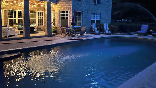 Una piscina con zona de patio, mobiliario exterior y un edificio con varias ventanas y un balcón.