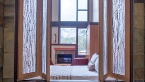 A bedroom with a fireplace, a bed, and a view through a window with a decorative frame. - Sunshine Mountain Lodge - Superior Room (Banff)