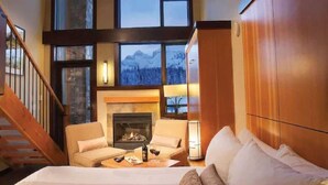 A cozy room with a fireplace, a sofa, a bed, and a view of mountains through large windows. - Sunshine Mountain Lodge - Superior Room (Banff)
