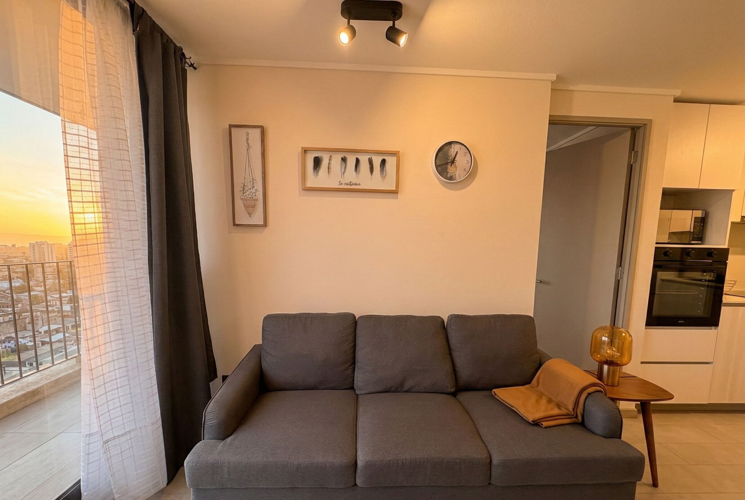 Una sala de estar moderna con un sofá gris, una mesita con una lámpara, una obra de arte enmarcada y un reloj en la pared.