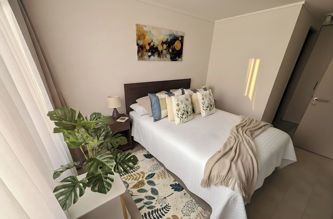 Ein ordentlich eingerichtetes Schlafzimmer mit Bett, Nachttisch, Lampe und einer großen Topfpflanze.