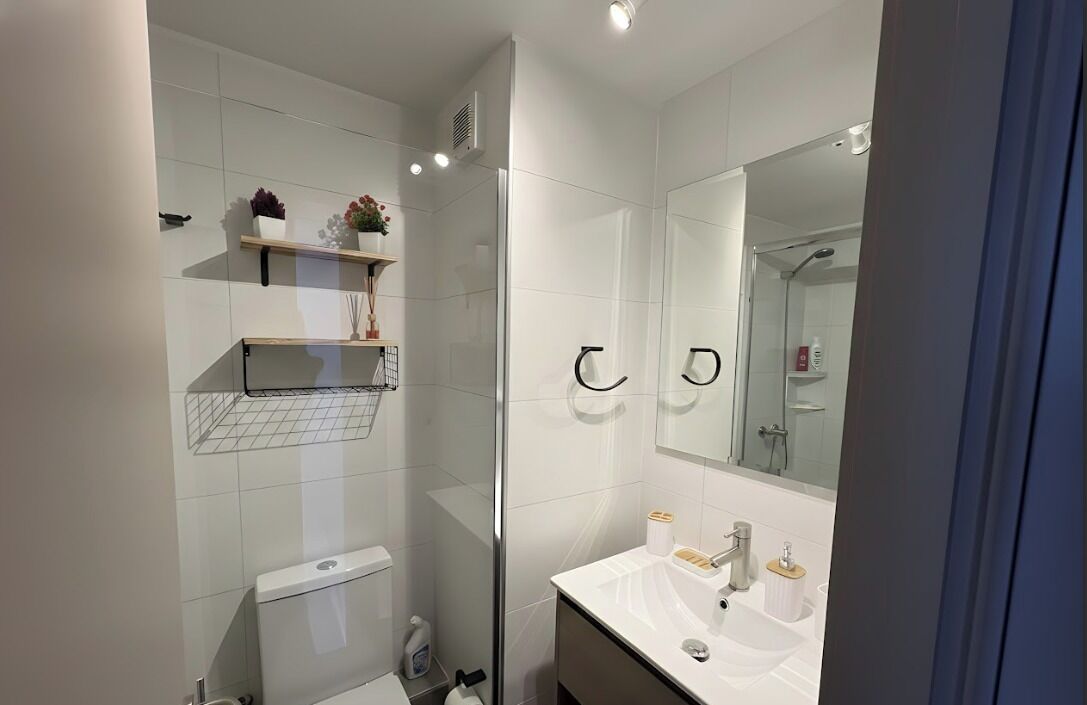 Un baño moderno con azulejos blancos, cabina de ducha de vidrio, tocador con espejo e inodoro.