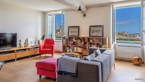 Un salon moderne avec une grande télévision, des canapés rouges et gris, une bibliothèque et une vue sur la mer.