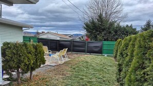 Un jardin avec une pelouse, un petit arbre, une terrasse avec des chaises et une clôture verte.